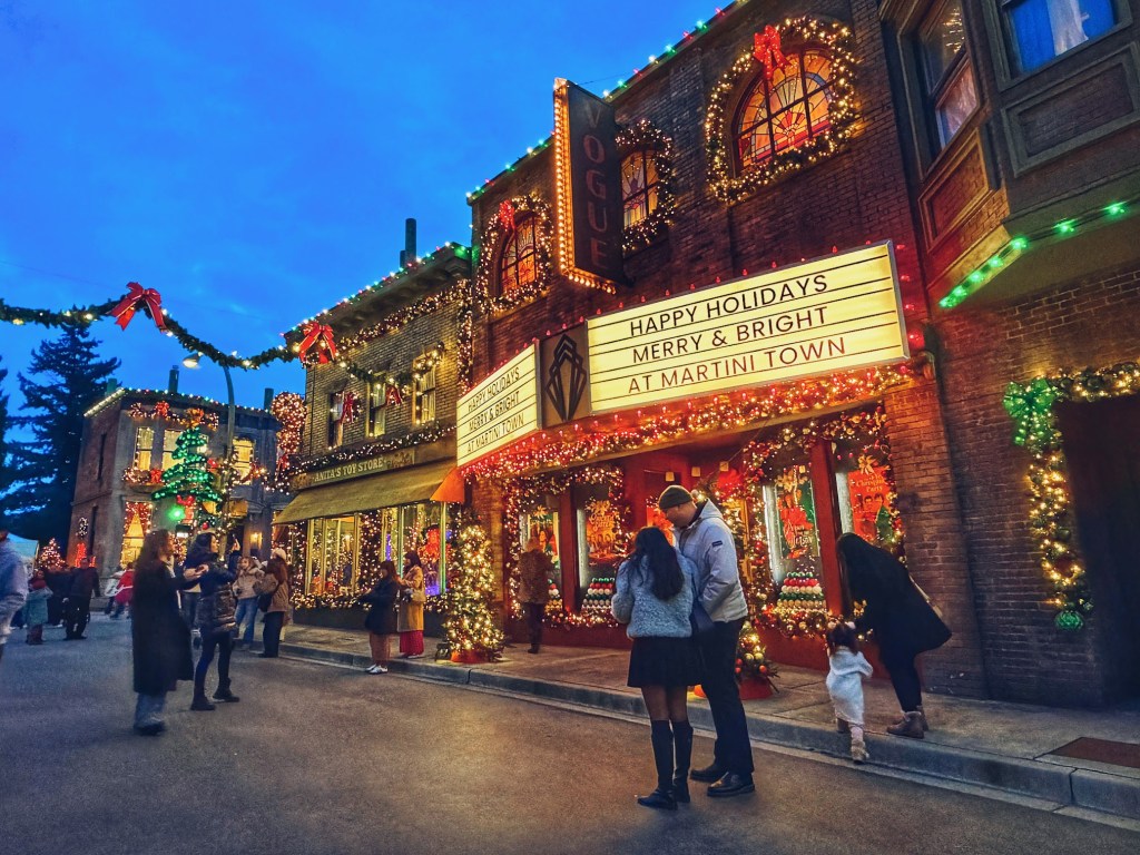 Visitors walking past the vintage Vogue Theater marquee at the Martini Town Merry & Bright holiday event, featuring brick buildings illuminated with warm Christmas lights, wreaths, and festive garlands under a twilight sky