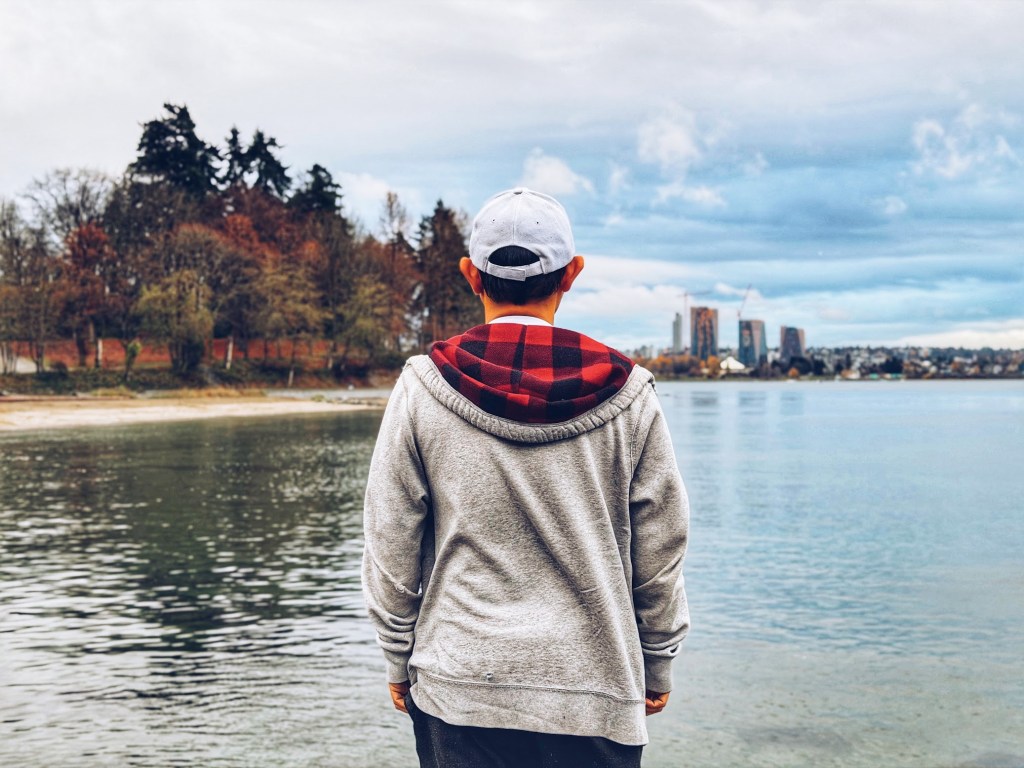 Young person in grey hoodie and white cap gazes at Vancouver skyline from Stanley Park Seawall waterfront with winter trees and sandy shore under overcast sky.